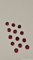 Photo shows 13 small, round-cut natural rubies with deep vivid electric red color and high clarity, displayed on plain background.