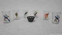 Front view of six crystal tumblers each decorated with colorful exotic bird illustrations, arranged in a row with the miniature bowl in the center.