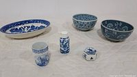 Seven blue and white porcelain items arranged on a quilted surface including oval platter, bowls, small vase, pepper shaker, trinket box, and cup.