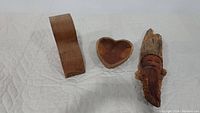 Wide view of all three carved wood items on fabric surface: corbel, heart-shaped bowl, and Old Man Face sculpture