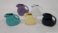 Five Fiesta Ware pitchers displayed together: two large and three smaller pitchers in purple, white, teal, yellow, and navy blue colors.