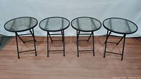 Full view of four round glass top folding patio tables arranged side by side on wooden floor.