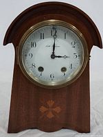 Front view of the clock showing wooden case with decorative inlay and clock face with black numerals and hands.