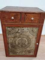 Front view of old wooden cabinet showing punched tin front door and two drawers with white knobs.