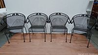 Front view of all four black wrought iron barrel patio chairs in a row, showing curved backs and mesh seating