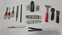 All tools arranged in a single row on a light background showing chisel handles, files, plane tool, utility knife, wire stripper, scrapers, and cutting tools