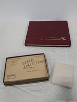 Reader's Digest maroon binder labeled 'First Day Cover Collection' alongside a box labeled 'Stamps Collection' and a small white envelope on white surface.