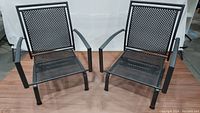 Pair of black Kettler Reno wrought iron patio arm chairs with lattice seats and backs shown side by side on wood flooring against a white backdrop.