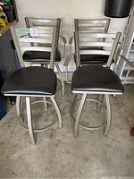 Four silver-tone metal bar stools with black cushioned seats and ladder-back designs photographed from the front in a garage.