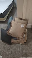 View of throw pillows stacked on carpeting near bed frame and mattress, showing beige pillows with fringe and blue-gray and dark gray pillows.