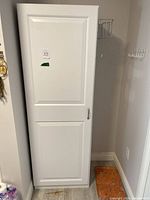 Closed white cabinet with panel door and handle, standing next to wall hooks and a floor tile.
