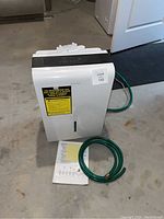 Dehumidifier, hose, and manual on basement floor from front-left angle