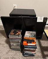 Dynex TV screen and stacks of CDs and DVDs in cases along with Firestick boxes on carpeted floor.