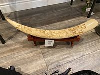 Faux elephant tusk carved with detailed dragon and ornamental carvings, displayed horizontally on wooden pedestal, showcasing the shape and carving detail as well as a visible crack on the tusk.