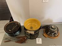 Overall view of fondue pot on burner, eight black plates stacked, and wooden-handled fondue forks spread on the table.