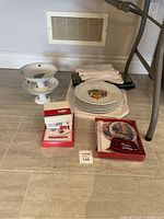 Photo showing two white footed candy dishes, stack of white napkins, six crest plates, holiday cards in boxes, and holiday coasters