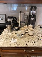 Photo showing three stainless steel travel mugs, Hamilton Beach kettle, Magic Bullet blender jar, SodaStream machine, and a group of ceramic mugs including Denby and souvenir mugs along with paper towel holder.