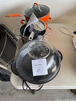 Stacked pots including an orange pot with glass lid and a metal splatter screen on top, displayed on a table.