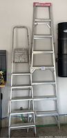 Two aluminum ladders, one with 8 steps and one with 5 steps, leaning against a white wall showing front view.