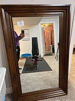 Full view of the large rectangular mirror with its dark wooden frame propped upright against a wall, showing room reflection and person holding phone.
