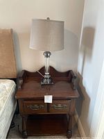 Wooden nightstand with two drawers, brass handles, and a lamp on top, showing overall appearance and size.