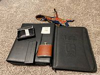 All items together on carpet: large black folder with embossed '100th Anniversary' logo, black Tahari wallet box, small hip flask with brown leather cover, black card holder box by Safari, multi-colored silk tie with paisley pattern.