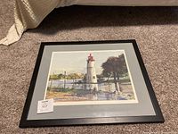 Framed picture shows the entire front view of the lighthouse picture with gray mat and black frame on the floor.