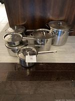 Six stainless steel pots of various sizes, stacked and arranged on the floor, each with matching lids featuring metal handles.