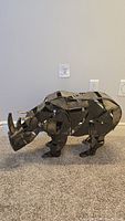 Left side view of metal rhinoceros sculpture showing its full body profile on carpeted floor near wall with electrical outlet.