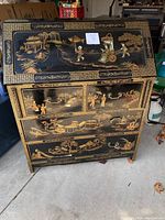 Front view of Oriental secretary desk showing black lacquer finish with gold detailed illustrations of landscapes and figures, with drawers and drop-front panel closed.