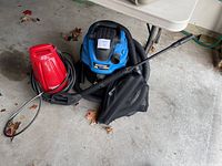 Red power washer and blue/black Mastervac shop vac with hose and collection bag beside a table on a concrete floor