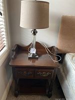 View of wooden nightstand showing two drawers with brass handles, lower shelf and table lamp on top with visible cord draped to the side.