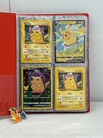 Four Pokémon cards featuring Pikachu from classic and recent series in a red binder with plastic sleeves, plus a Charizard pin on white background.