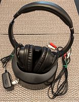 Front view of black wireless headphones on charging base, showing padded headband and ear cups with cables and charger placed beside them.