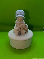Front angle photo showing small round white covered dish with girl sitting holding cat figurine on lid
