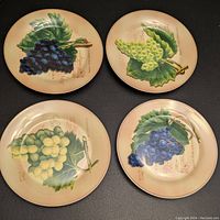 Four round porcelain dessert plates arranged on black background, each decorated with different grape cluster prints and vine leaves in vintage style with wine-related script text.