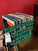 Green plastic crate filled with vinyl records against a red wall, showing the full quantity of albums in the crate.