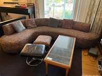 Full view of curved floral one-piece couch with five floral pillows and two gray pillows, placed on a black rug near window and furniture.