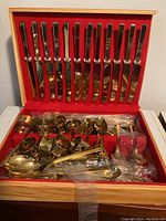 Open wooden cutlery box showing knives fixed on the lid and assorted brass cutlery items in the base, some wrapped in plastic.