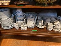 Photo shows assorted blue and white ceramic dishes neatly stacked on wooden shelves including plates, cups, saucers, and other serveware.