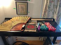 Large decorative wood and paper hand fan with intricate designs, alongside mats, candle holders, baseball bat, vacuum pump, and fabric coozies on black table.