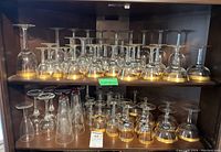 Photo showing multiple wine glasses on shelves, all with clear glass and gold patterned rim at base of bowl, various heights from 4 to 9 inches
