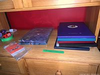 Eight stamp albums stacked on a wooden shelf, various colors including purple, blue, black, and patterned covers. Small cards indicating years and origin.