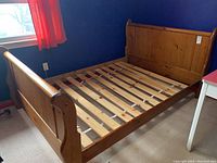 Wood sleigh bed frame in a room with blue walls and red curtain. Shows headboard, footboard, side rails, and wood slats for mattress support.