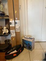 Photo showing two long cardboard boxes containing black tension rods next to a black and orange travel case and a smaller boxed item on floor.