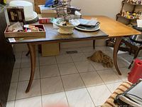 Wooden dining table with curved legs and two extension leaves, shown in room with tiled floor and various household items on top.