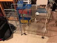 Two metal trolleys side by side on floor; larger gold-tone trolley with glass top and shelves on wheels, smaller silver metal wire trolley with wire basket shelves and wheels.