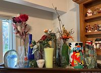 Wide view showing assortment of vases, paperweight, and faux flowers on shelf near window and cabinet with holiday decorations.