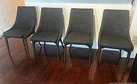 Four dark grey Structube dining chairs aligned in a row on a wooden floor against a white wall.