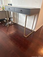 Full side and angled view of grey desk showing two drawers with ring pull handles and gold-toned metal geometric legs, placed on dark hardwood floor.